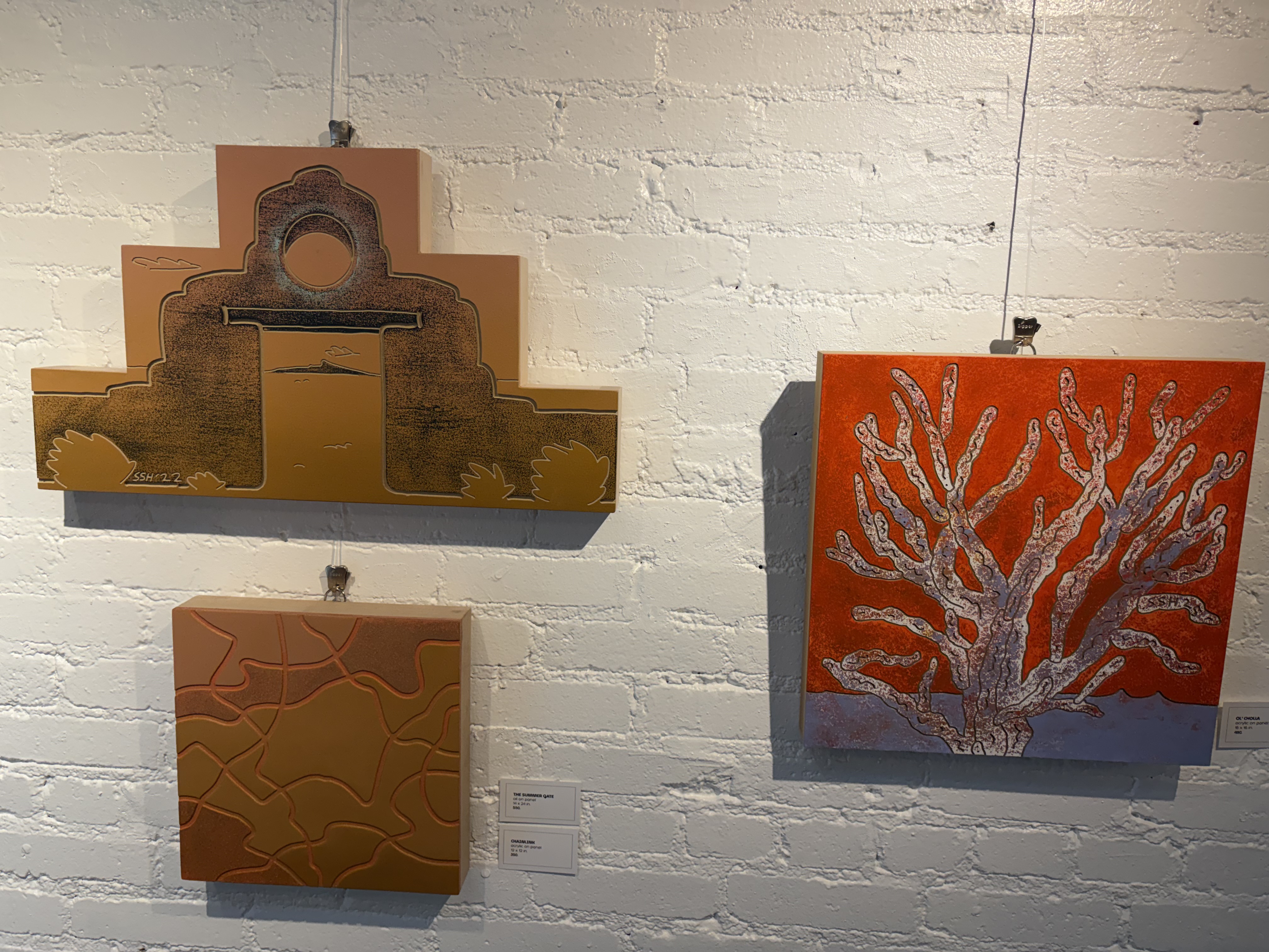 Three artworks on white painted brick: an adobe church form in ochre, a terracotta panel of curved lines, and a white tree on an orange ground.